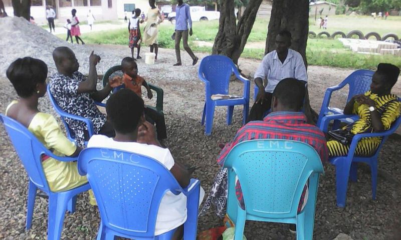 group of poeple sitting on plastic lawn chairs, under a tree, in a circle.