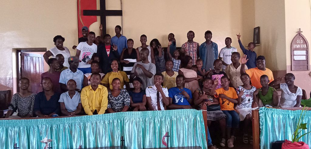 photo or a large group, seated in rows with the Methodist cross logo behind them, some signing I Love You, others waving.