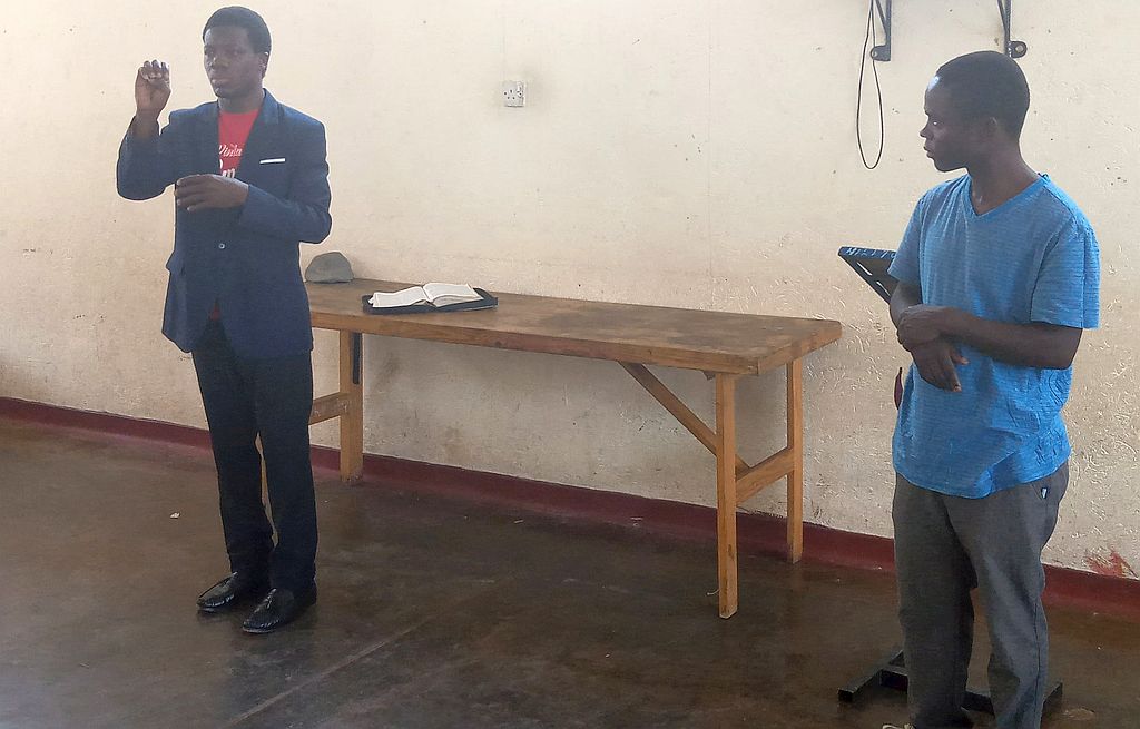 two men stand near a wall, signing, there is an open book behind them.